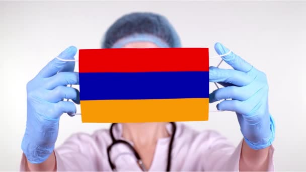 Primer plano. Doctor en gafas, gorra médica azul, guantes sostiene en las manos máscara médica con bandera de Armenia. Atención médica, protección por estado durante coronavirus, epidemia global. Concepto . — Vídeos de Stock