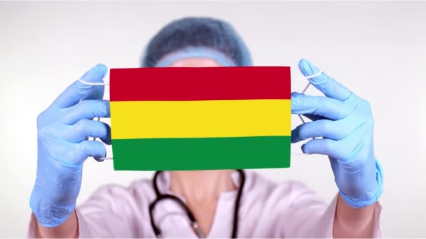 Zbliżenie. Lekarz w okularach, niebieska czapka medyczna, rękawiczki w rękach maska medyczna z flagą Boliwii. Opieka lekarzy, ochrona przez stan podczas koronawirusów, globalna epidemia. Pojęcie. — Wideo stockowe