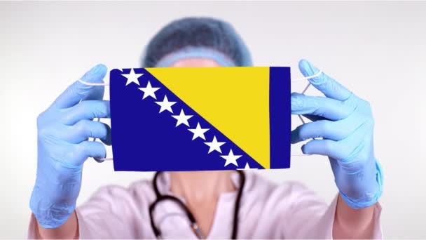 Yakın plan. Mavi şapkalı doktor, eldivenler Bosna-Hersek bayrağıyla el ele tıbbi maske tutuyor. Doktorlar, koronavirüs sırasında devletin koruması, küresel salgın. Kavram. — Stok video