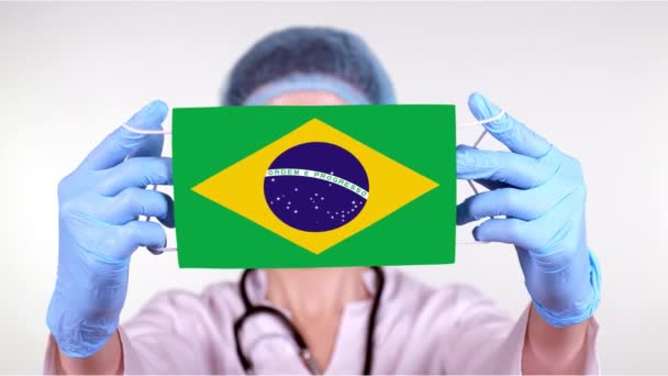 Close-up. Doutor em óculos, boné médico azul, luvas segura em mãos máscara médica com bandeira do Brasil. Cuidados médicos, proteção por estado durante o coronavírus, epidemia global. Conceito . — Vídeo de Stock