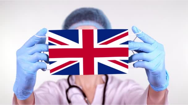 Close-up. Dokter in bril, blauwe medische pet, handschoenen houdt in handen medisch masker met vlag Verenigd Koninkrijk. Artsen zorgen, bescherming door de staat tijdens het coronavirus, wereldwijde epidemie. Concept. — Stockvideo