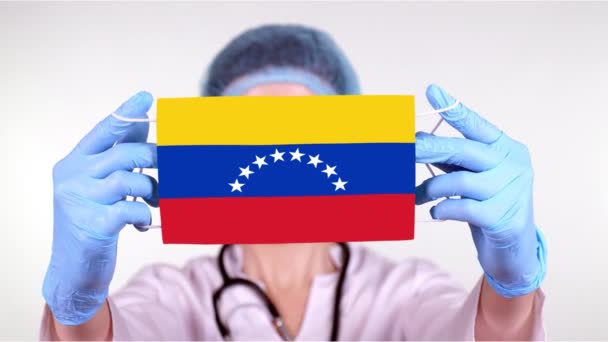 Primer plano. Doctor en gafas, gorra médica azul, guantes sostiene en las manos máscara médica con bandera de Venezuela. Atención médica, protección por estado durante coronavirus, epidemia global. Concepto . — Vídeos de Stock