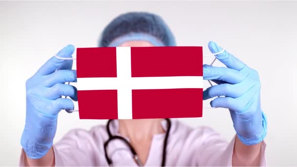 Gros plan. Docteur en lunettes, casquette médicale bleue, gants tient dans les mains masque médical avec drapeau du Danemark. Soins médicaux, protection par l'État pendant le coronavirus, épidémie mondiale. Concept . — Video