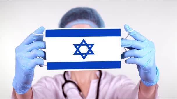 Primer plano. Doctor en gafas, gorra médica azul, guantes sostiene en las manos máscara médica con bandera de Israel. Atención médica, protección por estado durante coronavirus, epidemia global. Concepto . — Vídeo de stock