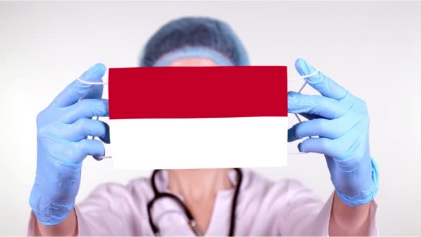 Primer plano. Doctor en gafas, gorra médica azul, guantes sostiene en las manos máscara médica con bandera de Indonesia. Atención médica, protección por estado durante coronavirus, epidemia global. Concepto . — Vídeos de Stock