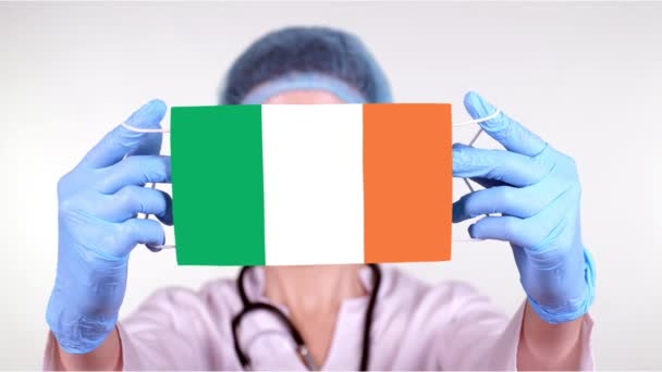 Primer plano. Doctor en gafas, gorra médica azul, guantes sostiene en las manos máscara médica con bandera de Irlanda. Atención médica, protección por estado durante coronavirus, epidemia global. Concepto . — Vídeos de Stock