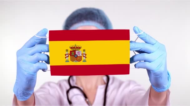 Yakın plan. Gözlüklü doktor, mavi tıbbi şapkalı, eldivenleri elinde İspanya bayrağıyla tıbbi maske tutuyor. Doktorlar, koronavirüs sırasında devletin koruması, küresel salgın. Kavram. — Stok video