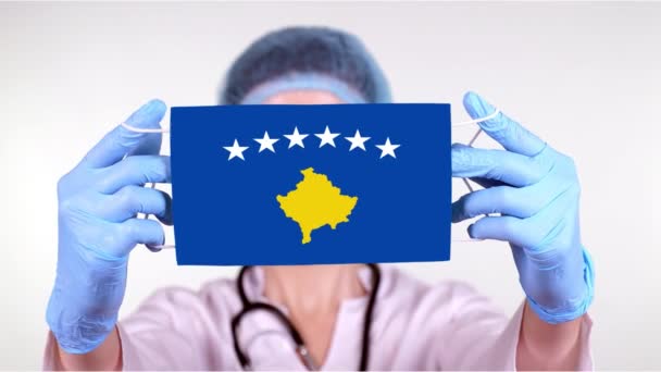 Nahaufnahme. Arzt mit Brille, blauer Mütze, Handschuhen hält medizinische Maske mit Kosovo-Fahne in den Händen. Ärzte kümmern sich, staatlicher Schutz beim Coronavirus, globale Epidemie. Konzept. — Stockvideo