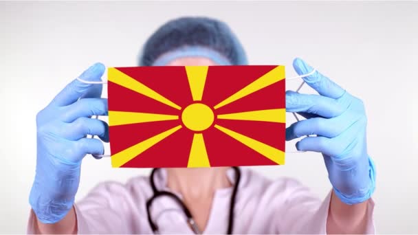 Yakın plan. Gözlüklü doktor, mavi tıbbi şapkalı, eldivenli Makedonya bayrağıyla el maskesi takıyor. Doktorlar, koronavirüs sırasında devletin koruması, küresel salgın. Kavram. — Stok video