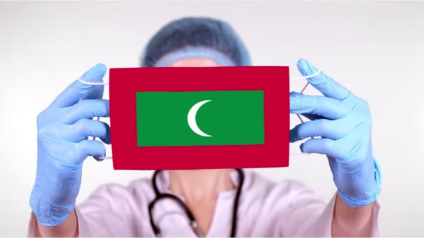 Close-up. Doctor in glasses, blue medical cap, gloves holds in hands medical mask with Maldives flag. Physicians care, protection by state during coronavirus, global epidemic. Concept. — Stock Video