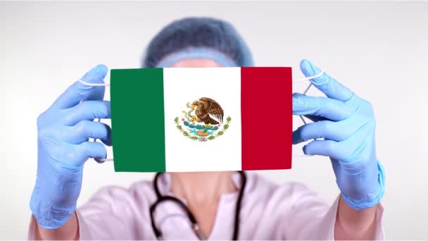 Primer plano. Doctor en gafas, gorra médica azul, guantes sostiene en las manos máscara médica con bandera de México. Atención médica, protección por estado durante coronavirus, epidemia global. Concepto . — Vídeos de Stock