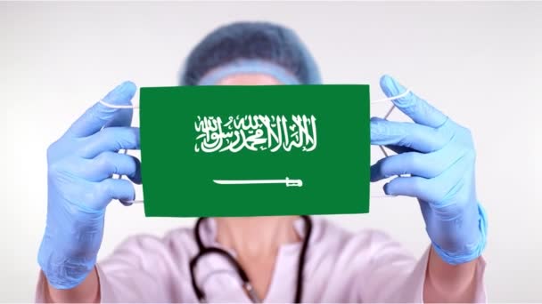 Primer plano. Doctor en gafas, gorra médica azul, guantes sostiene en las manos máscara médica con bandera de Arabia Saudita. Atención médica, protección por estado durante coronavirus, epidemia global. Concepto . — Vídeos de Stock