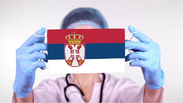 Yakın plan. Gözlüklü doktor, mavi tıbbi şapkalı, eldivenli Sırbistan bayrağıyla el maskesi takıyor. Doktorlar, koronavirüs sırasında devletin koruması, küresel salgın. Kavram. — Stok video
