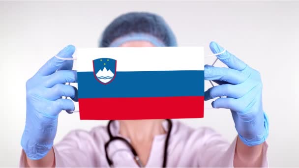 特写。戴眼镜的医生，戴着蓝色的医疗帽，戴着手套，手持印有斯洛文尼亚国旗的医疗面罩。医生关心，在验尸官出现时受到国家的保护，全球性的流行病。概念. — 图库视频影像