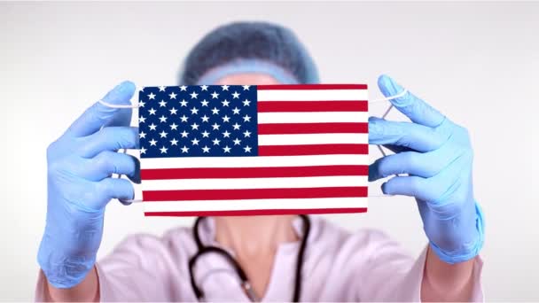 Κοντινό πλάνο. Γιατρός με γυαλιά, μπλε ιατρικό καπέλο, γάντια κρατούν στα χέρια ιατρική μάσκα με σημαία των ΗΠΑ. Οι γιατροί φροντίζουν, προστασία από το κράτος κατά τη διάρκεια του κορωναϊού, παγκόσμια επιδημία. Έννοια. — Αρχείο Βίντεο