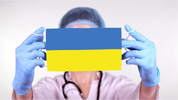 Detailní záběr. Doktor v brýlích, modrá lékařská čepice, rukavice drží v ruce lékařskou masku s vlajkou Ukrajiny. Lékaři pečují, chrání stát během koronaviru, globální epidemie. Koncept. — Stock video
