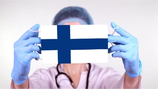 Gros plan. Docteur en lunettes, casquette médicale bleue, gants tient dans les mains masque médical avec drapeau de la Finlande. Soins médicaux, protection par l'État pendant le coronavirus, épidémie mondiale. Concept . — Video