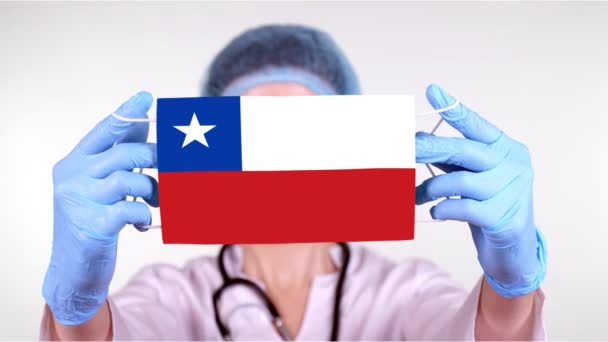 特写。戴眼镜的医生，戴着蓝色的医疗帽，戴着手套，手持印有智利国旗的医用口罩。医生关心，在验尸官出现时受到国家的保护，全球性的流行病。概念. — 图库视频影像