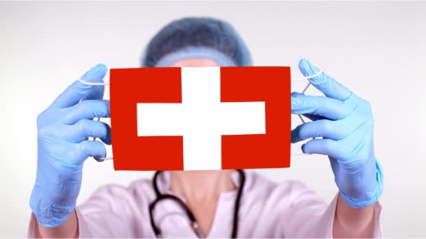 Yakın plan. Gözlüklü doktor, mavi tıbbi şapkalı, eldivenli, İsviçre bayrağıyla tıbbi maske takıyor. Doktorlar, koronavirüs sırasında devletin koruması, küresel salgın. Kavram. — Stok video