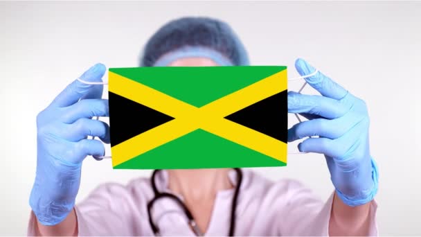 Primer plano. Doctor en gafas, gorra médica azul, guantes sostiene en las manos máscara médica con bandera de Jamaica. Atención médica, protección por estado durante coronavirus, epidemia global. Concepto . — Vídeos de Stock