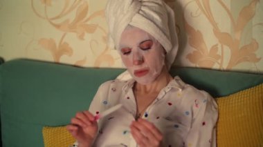 Portre, kozmetik kumaş maskeli bir kadın ve kafasında beyaz bir havluyla, tırnaklarını törpüyle törpülüyor, manikür yaptırıyor, geceleri, TV ekranının ışığının altında.,