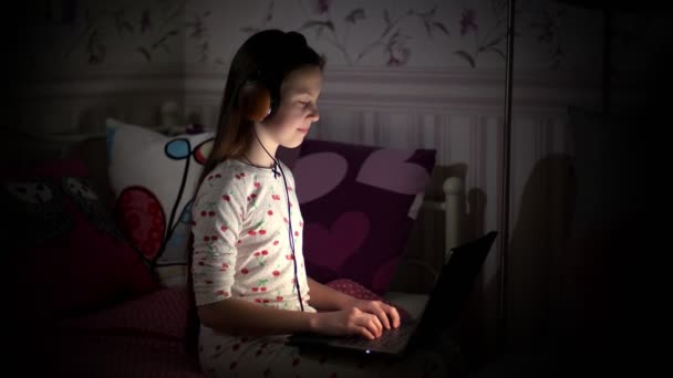 Dospívající dívka, v pyžamu, ve sluchátkách, dívá se na něco na notebooku, píše, poslouchá hudbu, v noci, v ložnici. dítě, teenager pracuje na notebooku v noci — Stock video