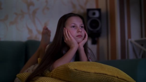 Дівчина-підліток у піжамі дивиться фільм по телевізору в темряві, лежить на дивані. Кімната освітлена світлом з екрану телевізора . — стокове відео