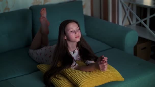 Adolescente en pijama viendo una película en la televisión en la oscuridad, acostada en el sofá. La habitación está iluminada por la luz de la pantalla del televisor . — Vídeo de stock