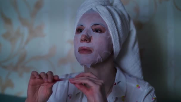 Portrét, žena v kosmetické látkové masce a s bílým ručníkem na hlavě, se v noci dívá na televizi a pilníkem na nehty si piluje nehty. — Stock video