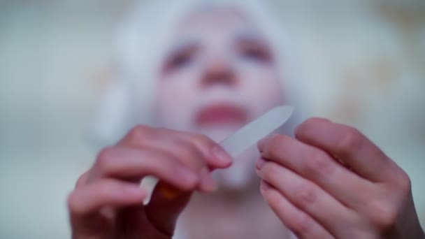 Primo piano, una donna in una maschera di tessuto cosmetico e con un asciugamano bianco sulla testa, si lima le unghie con una lima per unghie, manicure, di notte, sotto la luce dello schermo TV , — Video Stock