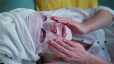 Portre, genç bayan, kozmetik kumaş maskeli kadın ve kafasında beyaz bir havluyla kanepede uzanmış, evde güzellik prosedürleri sırasında dinleniyor..