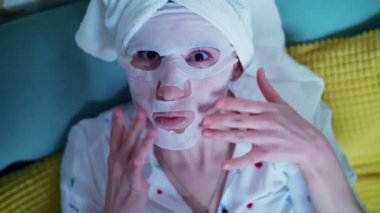 Portre, sevimli genç bayan, kozmetik kumaş maskeli kadın ve kafasında beyaz bir havluyla kanepede uzanmış, evde güzellik prosedürleri sırasında dinleniyor..