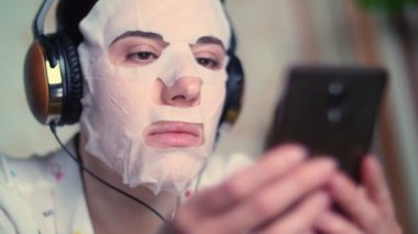 Portre, beyaz kozmetik kumaş maskeli, kulaklıklı, akıllı telefonlu, elinde cep telefonu olan genç bir kadın. Geceleri televizyon ekranının önünde, kanepede uzanıp onu dinliyor, izliyordum.