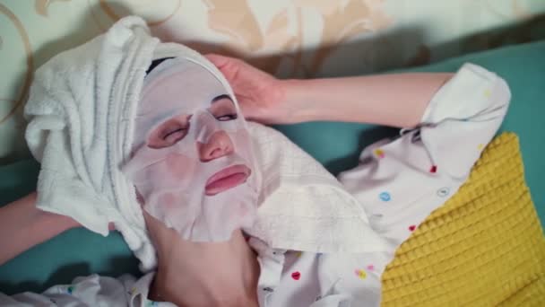 Портрет, женщина в маске из косметической ткани и с белым полотенцем на голове засыпает перед телевизором. ночь, свет с экрана телевизора . — стоковое видео