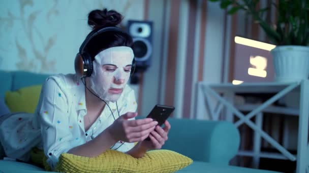 Portret, młoda kobieta w białej masce z tkaniny kosmetycznej, ze słuchawkami na głowie, trzyma smartfona, mobilny w rękach. słuchanie, oglądanie smth, w nocy, leżąc na sofie, przed ekranem telewizora — Wideo stockowe