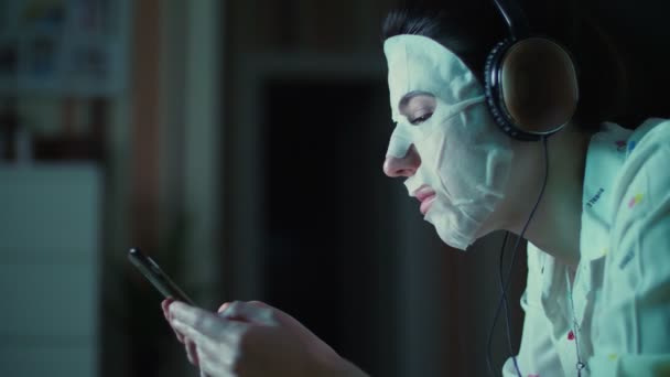 Портрет, молодая женщина в белой косметической маске, с наушниками на голове, держит смартфон, мобильный в руках. слушать, смотреть запах, ночью, лежа на диване, перед экраном телевизора — стоковое видео