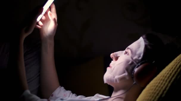 Donna in tessuto cosmetico bianco maschera, con cuffie, tiene smartphone, mobile in mano. ascoltare, guardare smth, di notte, sdraiato sul divano, davanti allo schermo — Video Stock