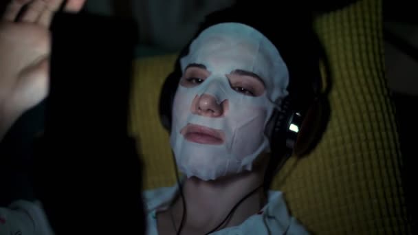 Mujer en máscara de tela cosmética blanca, con auriculares, sostiene el teléfono inteligente, móvil en las manos. escuchando, mirando smth, por la noche, acostado en el sofá, delante de la pantalla — Vídeo de stock
