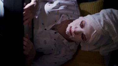 Kozmetik kumaş maskeli ve kafasında beyaz havluyla genç bir kadın geceleri dizüstü bilgisayarda çalışıyor, ekranda izliyor. Evde, kanepede çalışan yorgun iş kadını.