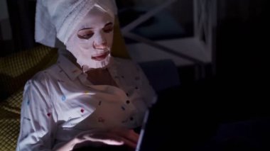 Kozmetik kumaş maskeli ve kafasında beyaz havluyla genç bir kadın geceleri dizüstü bilgisayarda çalışıyor, ekranda izliyor. Evde, kanepede çalışan yorgun iş kadını.