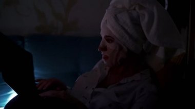 Kozmetik kumaş maskeli ve kafasında beyaz havluyla genç bir kadın geceleri dizüstü bilgisayarda çalışıyor, ekranda izliyor. Evde, kanepede çalışan yorgun iş kadını.