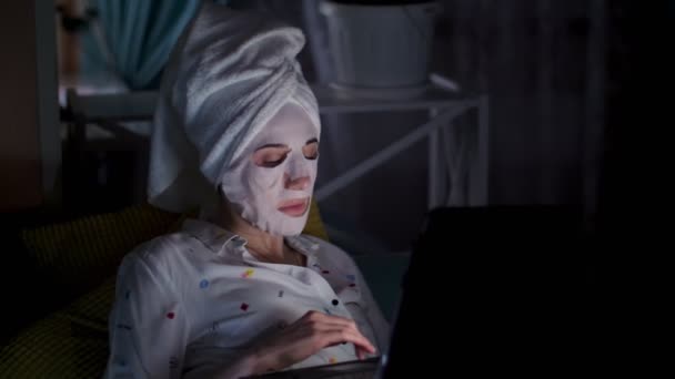 Ung kvinna i kosmetisk tyg mask och vit handduk på huvudet, arbetar på laptop, tittar på skärmen, på natten. trött affärskvinna arbetar hemma på soffan — Stockvideo