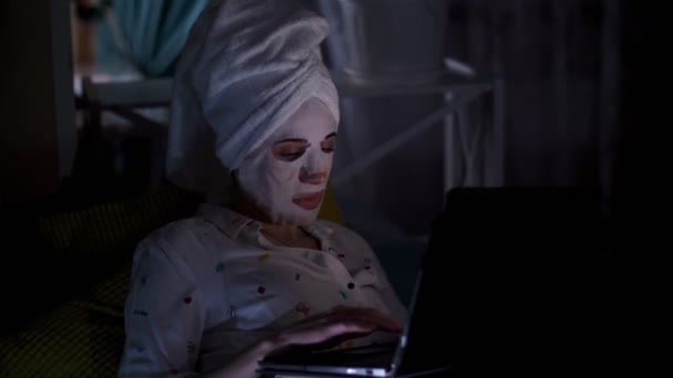 Νεαρή γυναίκα με μάσκα καλλυντικού υφάσματος και λευκή πετσέτα στο κεφάλι της, εργάζεται σε φορητό υπολογιστή, βλέποντας στην οθόνη, τη νύχτα. κουρασμένη επιχειρηματίας που εργάζεται στο σπίτι στον καναπέ — Αρχείο Βίντεο