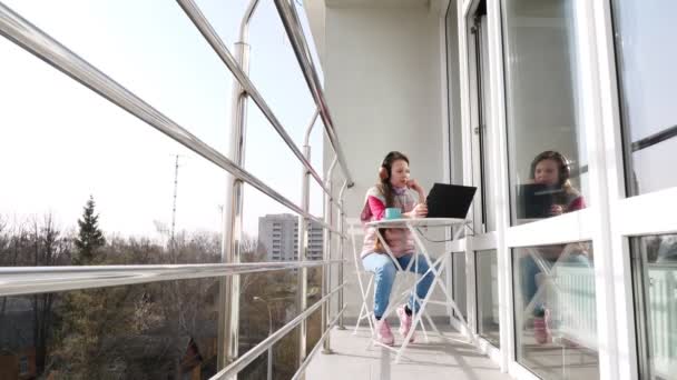 Kulaklıklı genç bir kız, dizüstü bilgisayarda çalışıyor, açık balkonda sohbet ediyor. Bahar güneşli bir gün. Karantina sırasında uzaktan öğrenme. Kal, evde çalış. Koronavirüs salgını. — Stok video