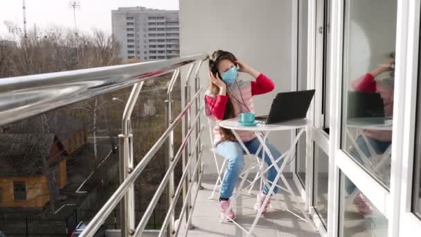 マスクとヘッドフォンのティーンエイジャーの女の子は、ノートパソコンで音楽を聞いて、オンラインでチャットし、オープンバルコニーで。春の晴れた日。隔離だ。家で勉強しろ。コロナウイルスの流行. — ストック動画