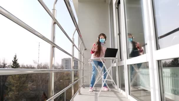Tiener meisje met masker en koptelefoon, luisteren naar muziek op laptop, chatten online, op een open balkon. Zonnige lentedag. quarantaine. Blijf, studeer thuis. Coronavirusepidemie. — Stockvideo