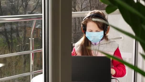 Tiener meisje met masker en koptelefoon, typen, werken op laptop, op een open balkon. Zonnige lentedag. afstandsonderwijs tijdens quarantaine. Blijf, studeer thuis. Coronavirusepidemie. — Stockvideo