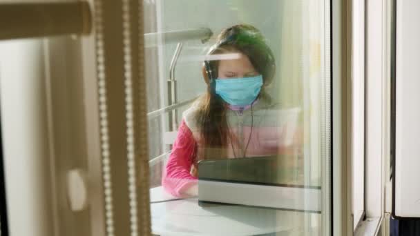 Maskeli ve kulaklıklı genç bir kız, yazı yazıyor, tablet üzerinde çalışıyor, açık balkonda çalışıyor. Bahar güneşli bir gün. Karantina sırasında uzak mesafe öğrenimi. Kal, evde çalış. Koronavirüs salgını. — Stok video