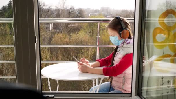 マスクとヘッドフォンのティーンエイジャーの女の子は、スマートフォンを使用して、オープンバルコニーで、自撮りになります。春の晴れた日。隔離の概念オンラインでチャット。家にいて。コロナウイルスの流行. — ストック動画