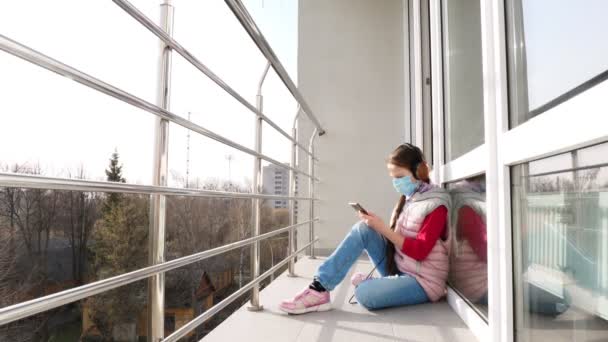 Chica adolescente en máscara y auriculares, escuchando música en el teléfono inteligente, en el balcón abierto. Primavera día soleado. cuarentena. Quédate, estudia en casa. epidemia de coronavirus . — Vídeos de Stock